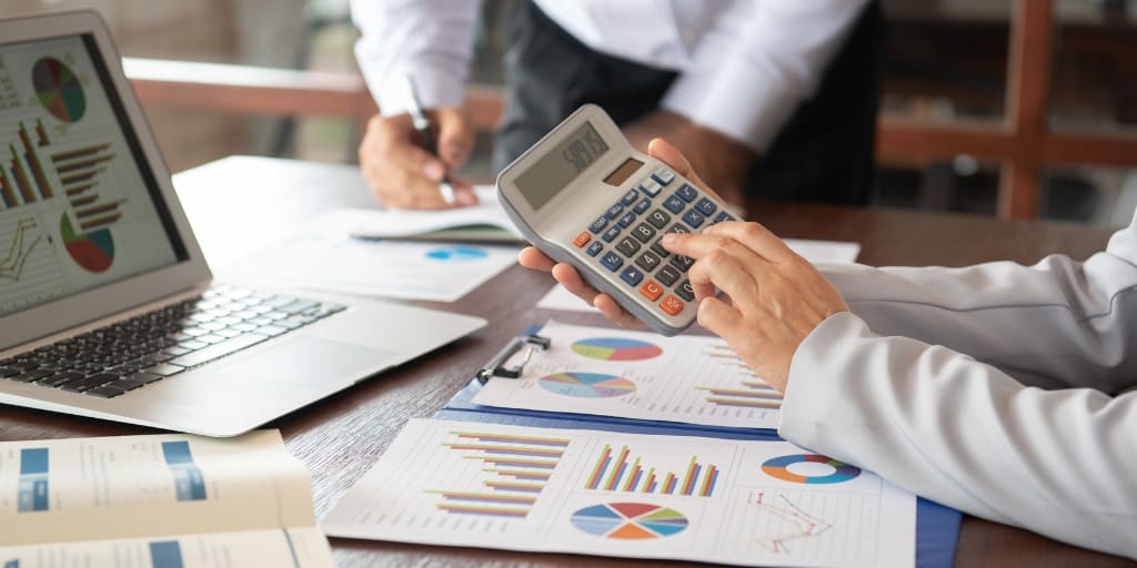Fotografía de una mesa de trabajo donde dos personas analizan información financiera. En primer plano, una persona utiliza una calculadora sobre documentos con gráficos e indicadores, mientras otra toma notas. También se observa un computador portátil con reportes financieros. La imagen ilustra el análisis financiero y acompaña la publicación del Decreto 0978 de 2026, relacionado con la financiación de micro, pequeñas y medianas empresas mediante procesos de titularización.